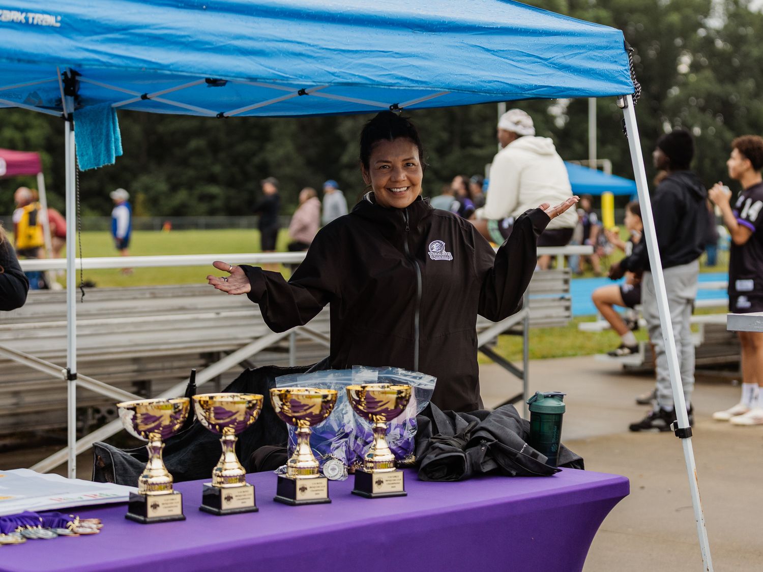 Smiling official stands behind trophy display table under blue canopy at outdoor sports event.