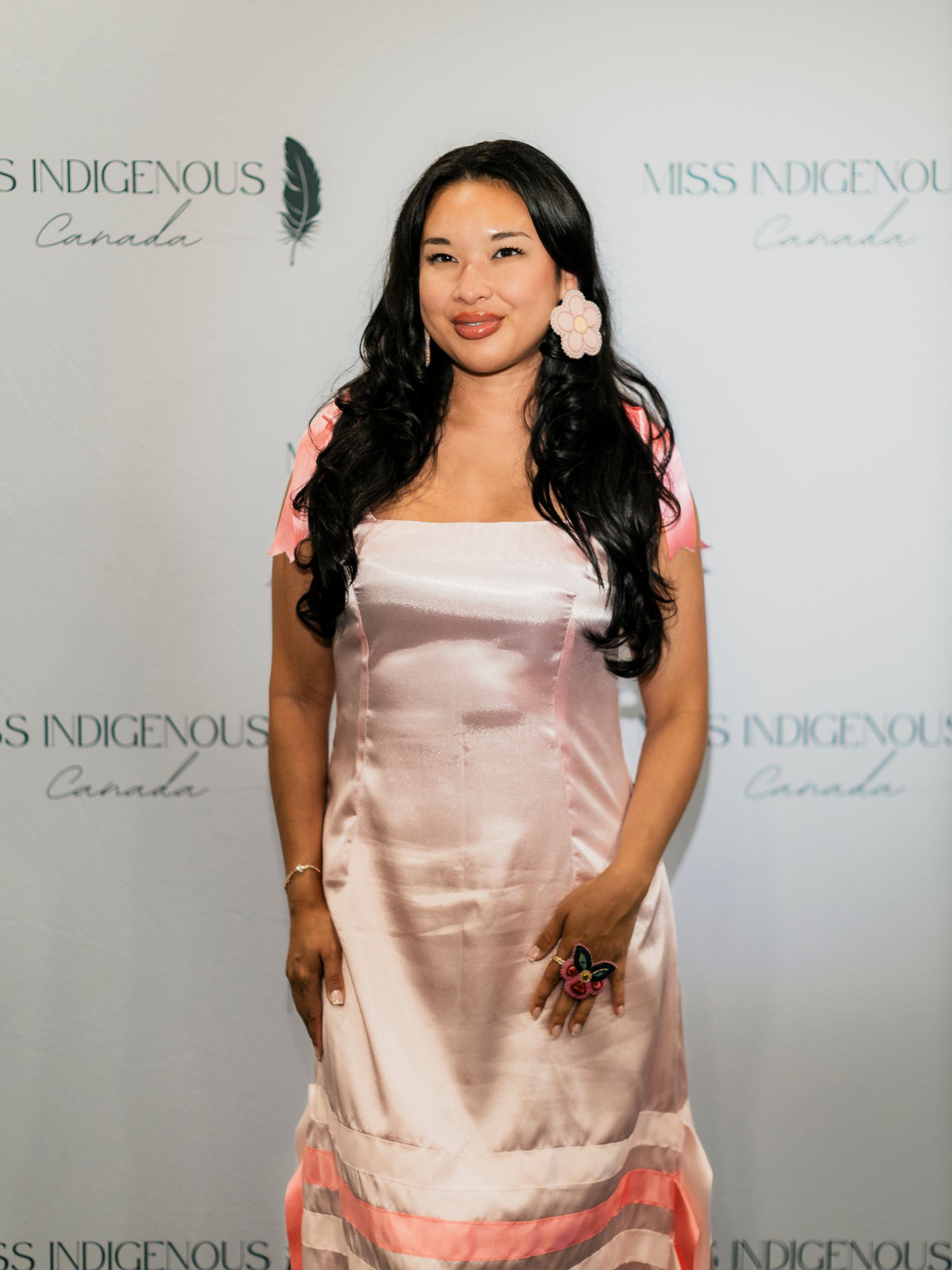 Woman in pink satin dress poses at Miss Indigenous Canada event backdrop.