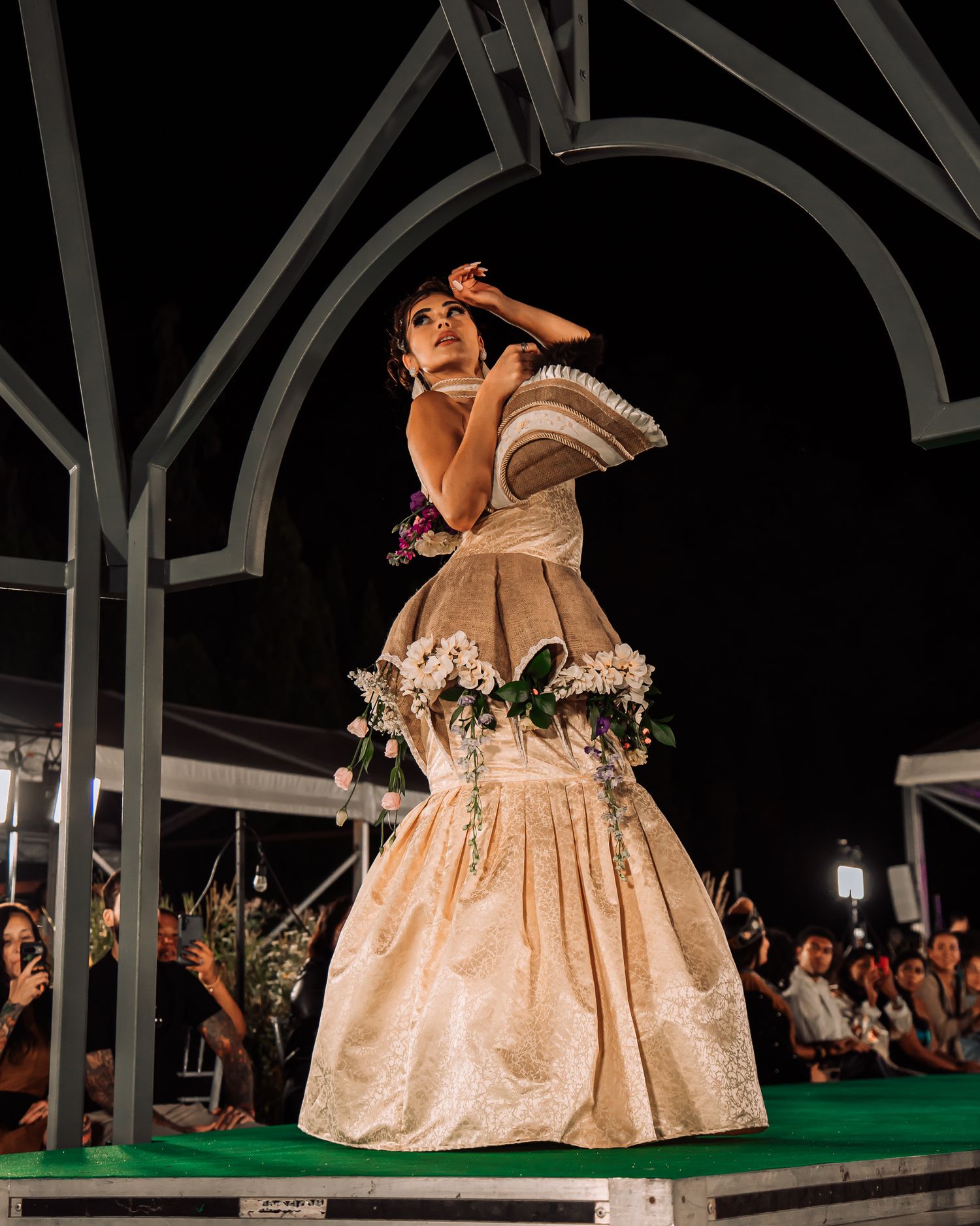Model in ornate cream ballgown with floral accents posing dramatically on stage at a nighttime fashion show.