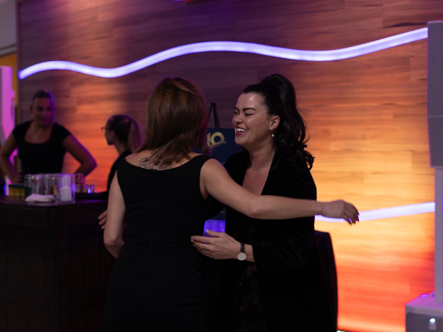 Two women in black dresses share a warm embrace at a stylish evening event with ambient lighting.