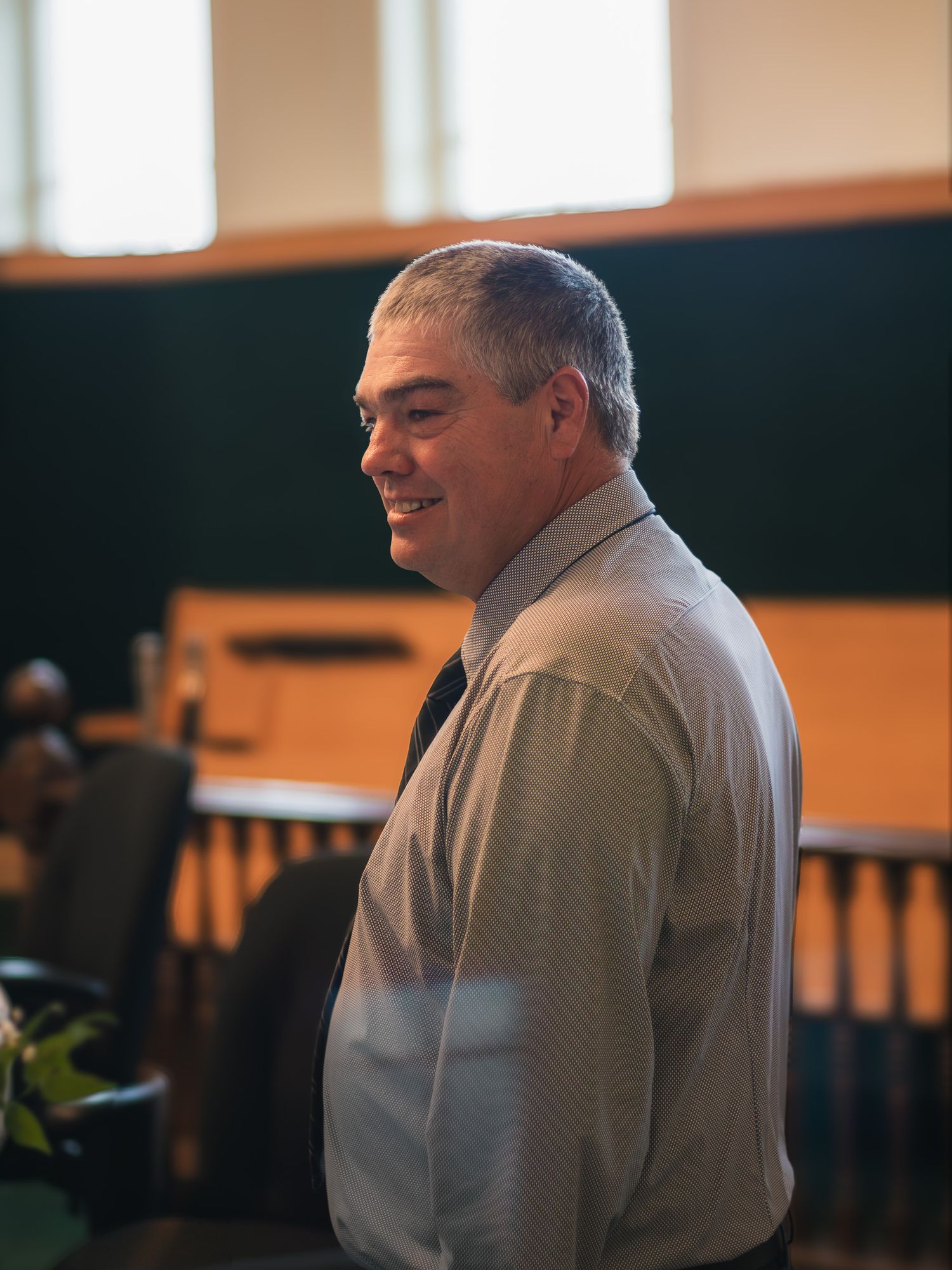 Smiling middle-aged man in gray dress shirt stands in elegant wood-paneled venue.