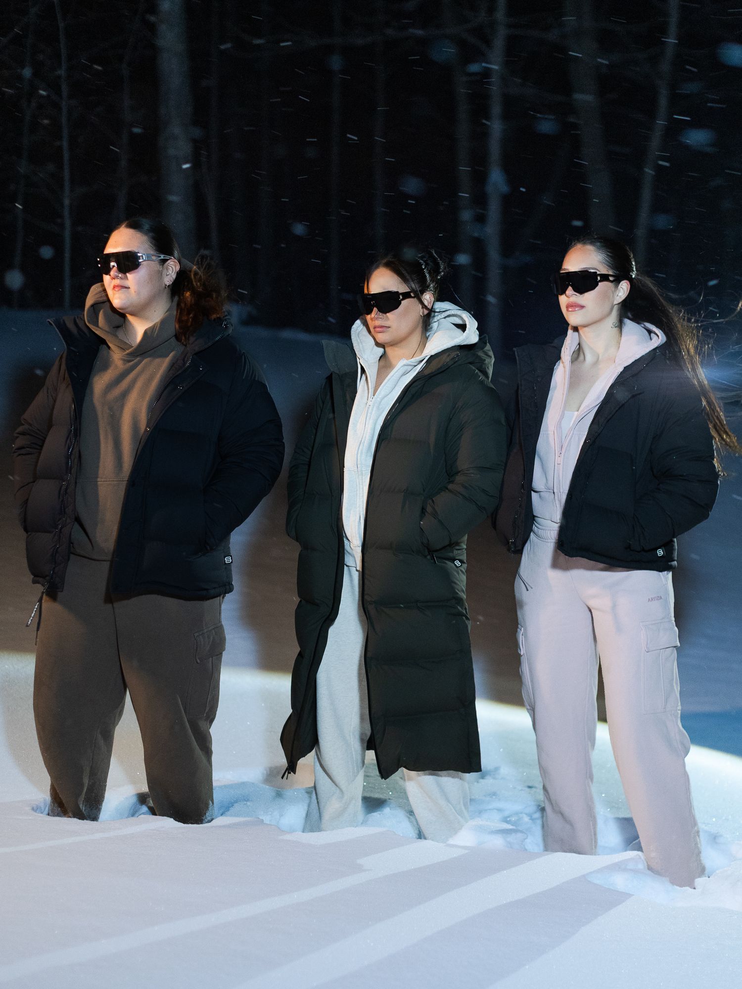 Three women in puffer coats and sunglasses stand in snowy winter landscape with dark forest behind.