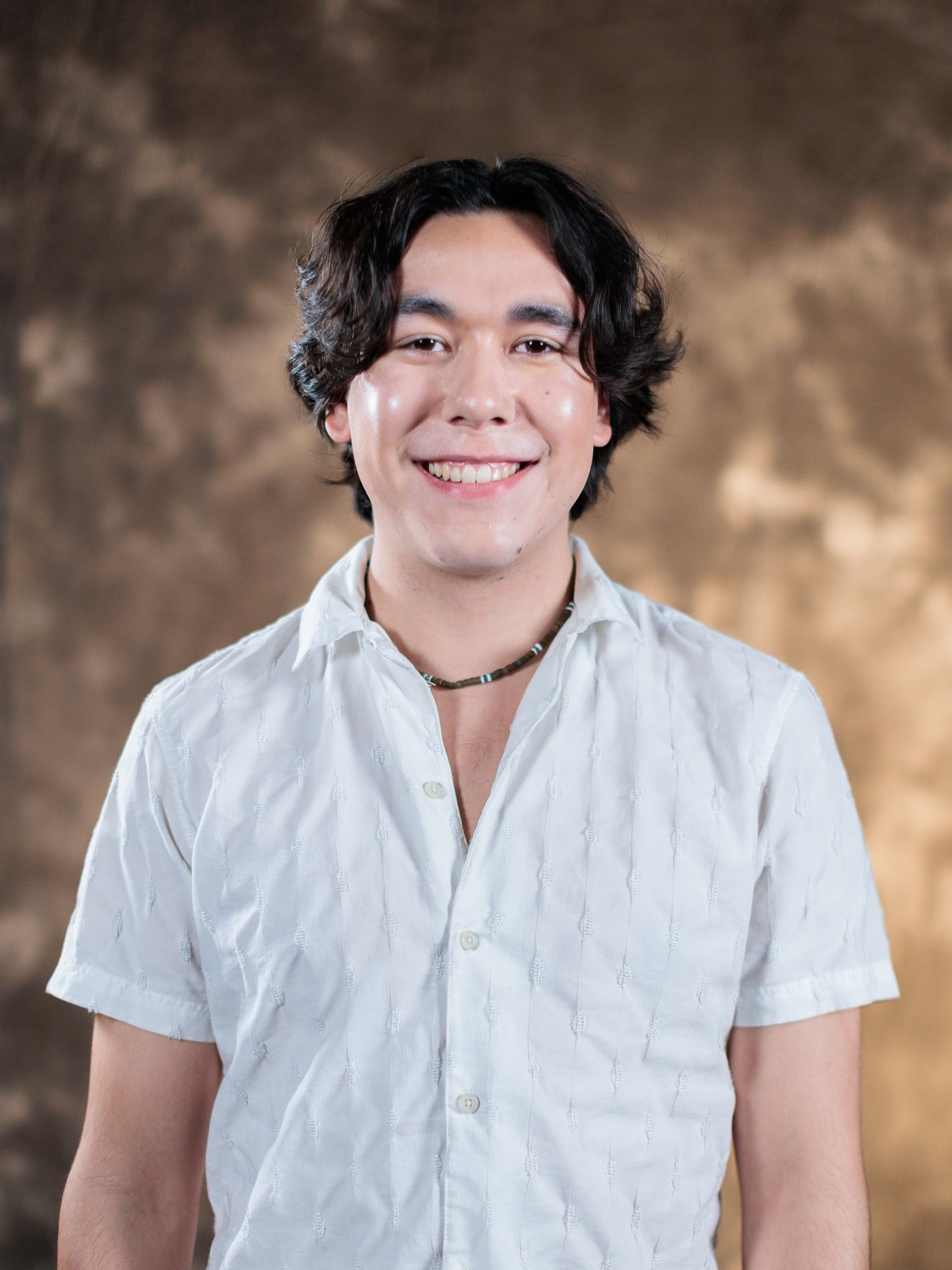 Smiling young man with dark wavy hair wearing a white button-up shirt and necklace against a brown backdrop.
