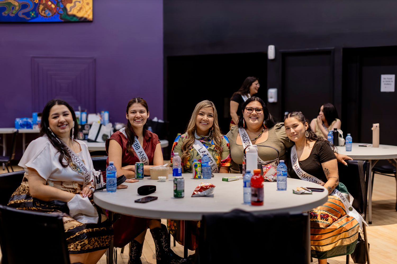 Group of five smiling women sitting together at a round table at an indoor social event.