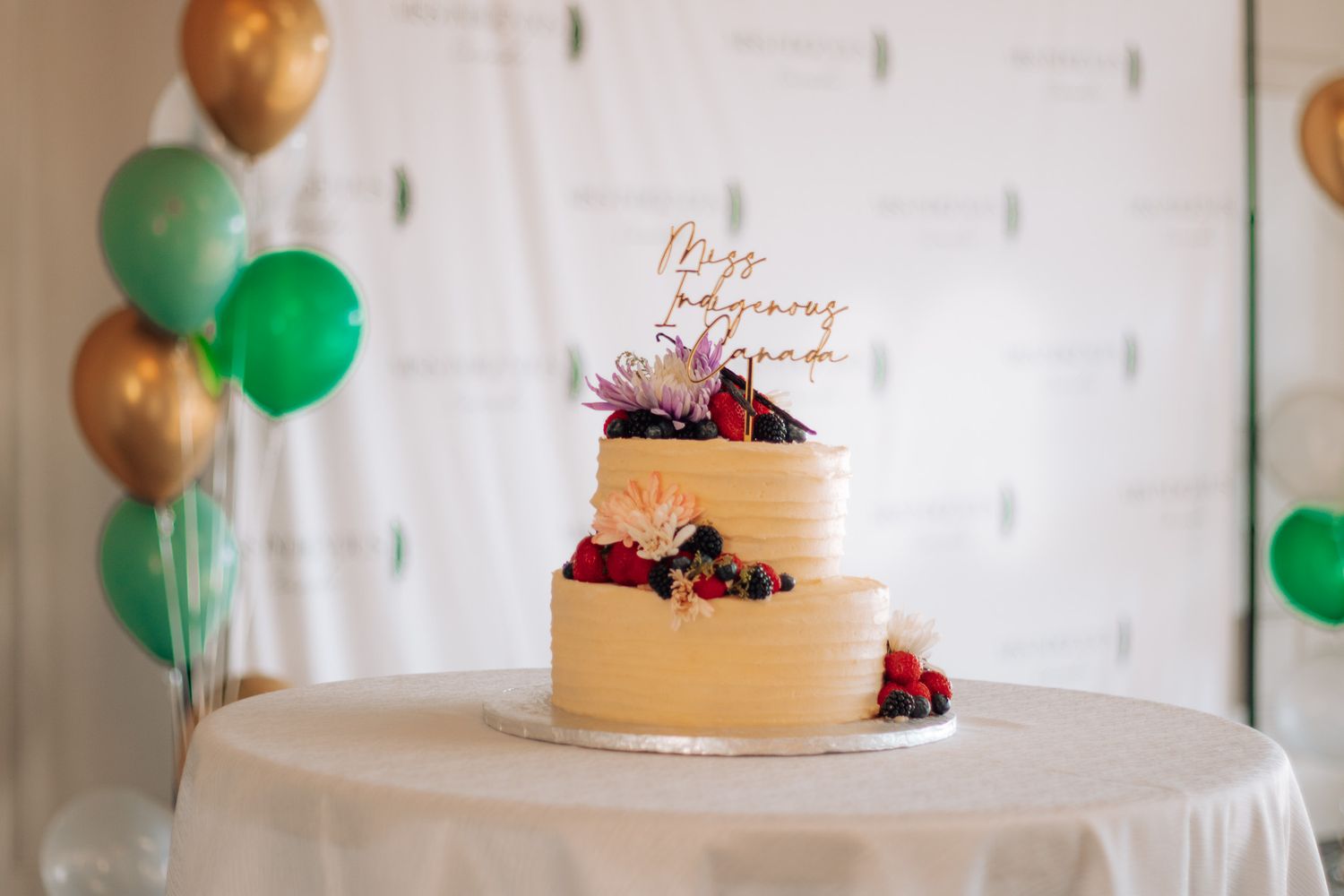 Elegant cake with berries displayed on white table with balloons in background.