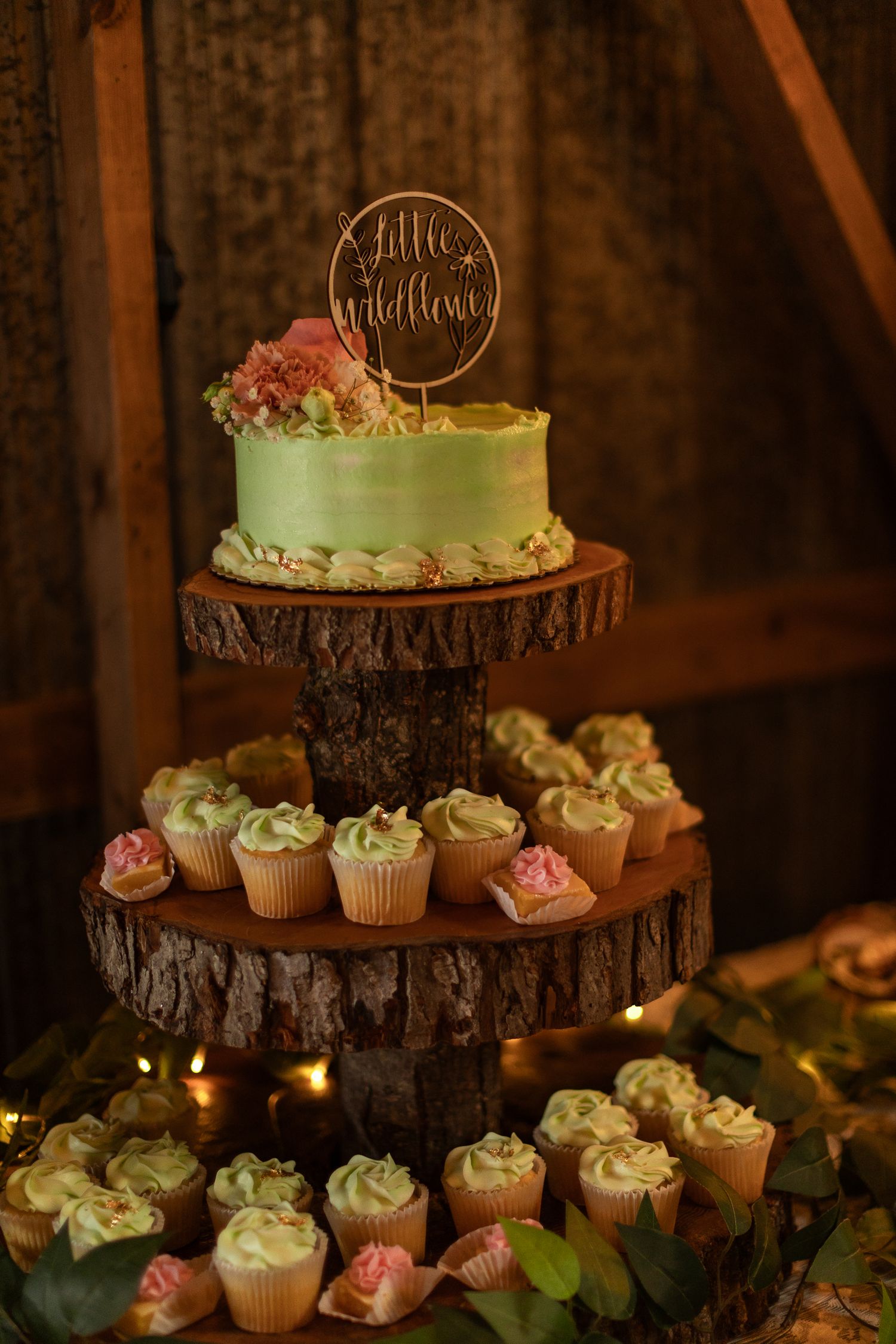 A rustic wedding cake display with a mint green cake and cupcakes arranged on wooden log slices with twinkle lights.