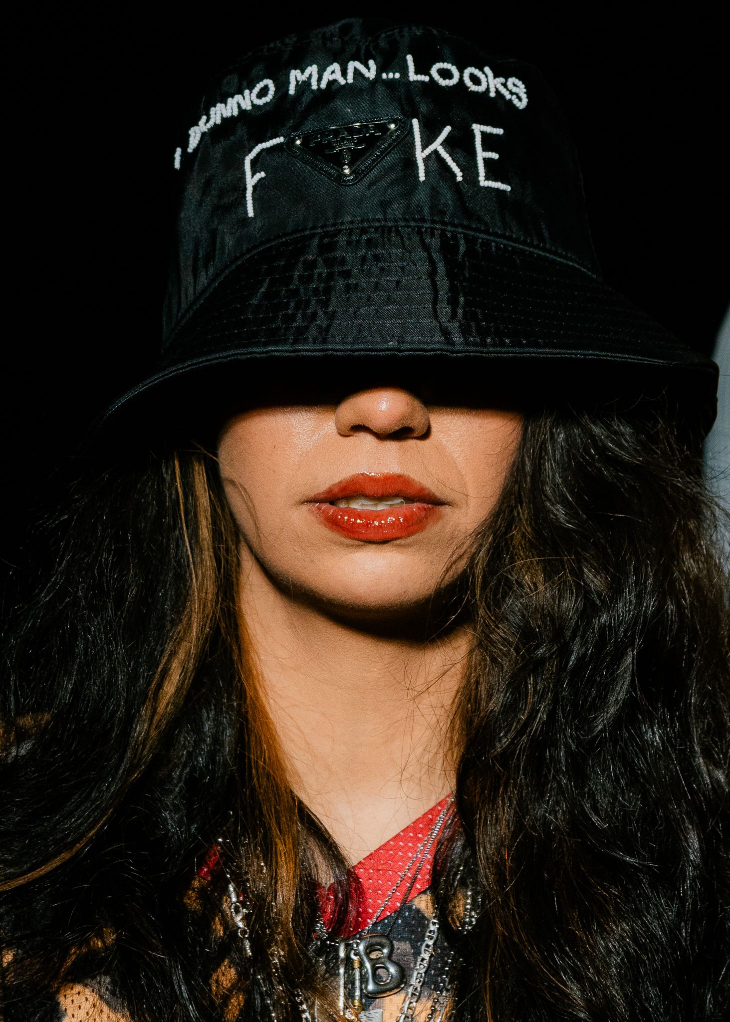 Woman wearing black bucket hat with FAKE text, long dark hair, orange lips, dark background.