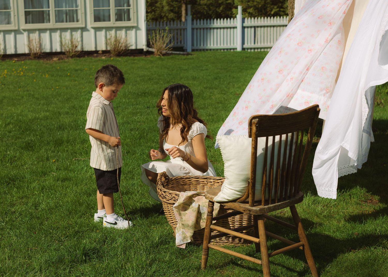 Woman sitting in wooden chair on green lawn folding laundry while young boy stands nearby beside hanging white sheets.