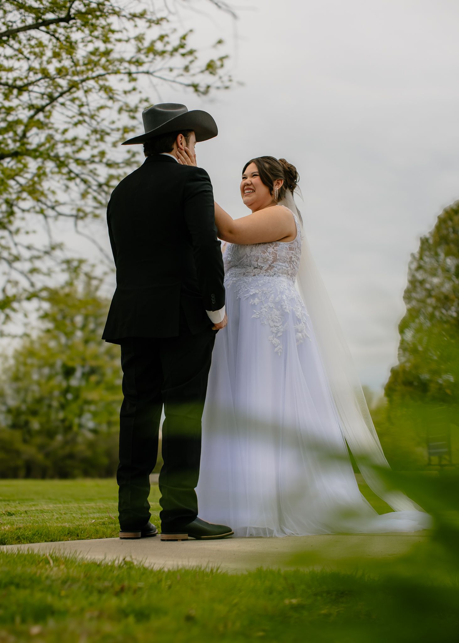Bride in white lace gown and groom in black suit with cowboy hat share a romantic outdoor moment.