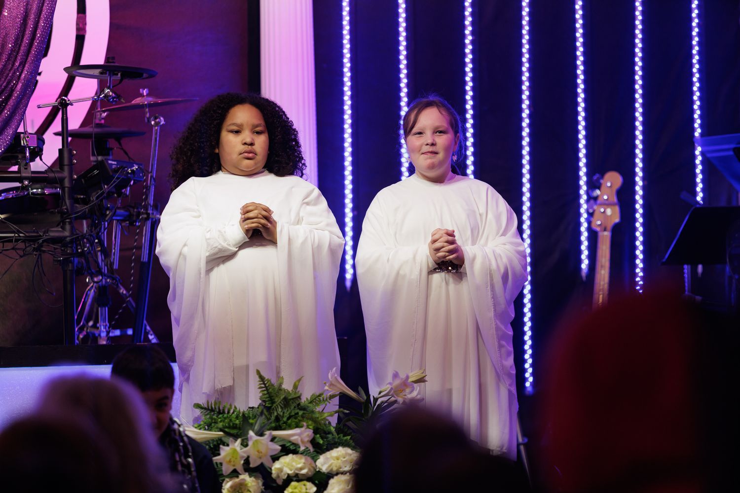 Two women in white robes stand on a stage with purple lights and flowers, appearing to perform at a formal event.