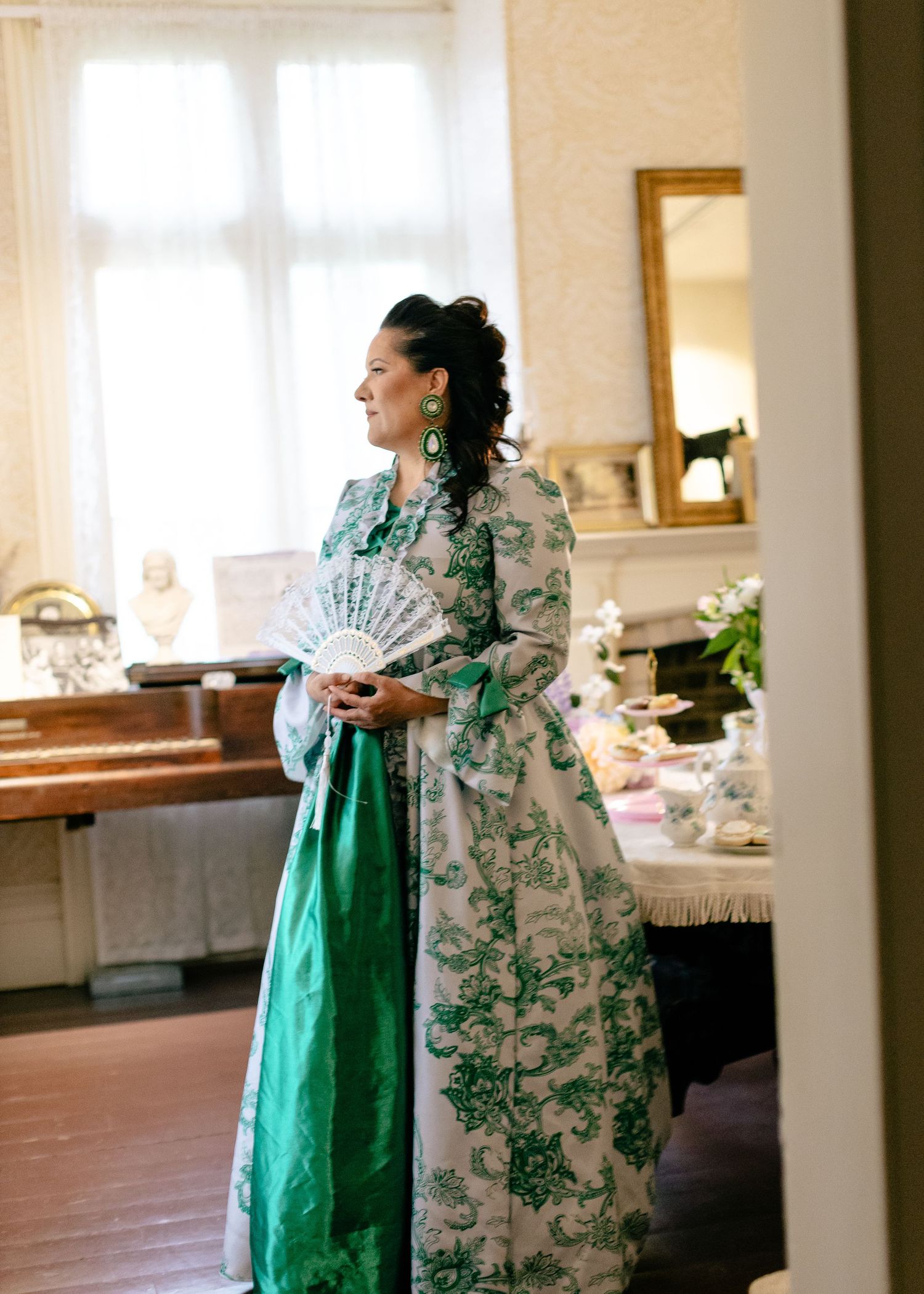 Woman in elegant green and white floral Victorian gown holding a fan standing in a vintage-styled room.