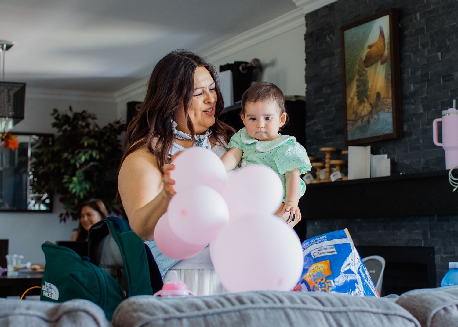 A mother holds her baby girl surrounded by pink balloons at a first birthday celebration indoors.