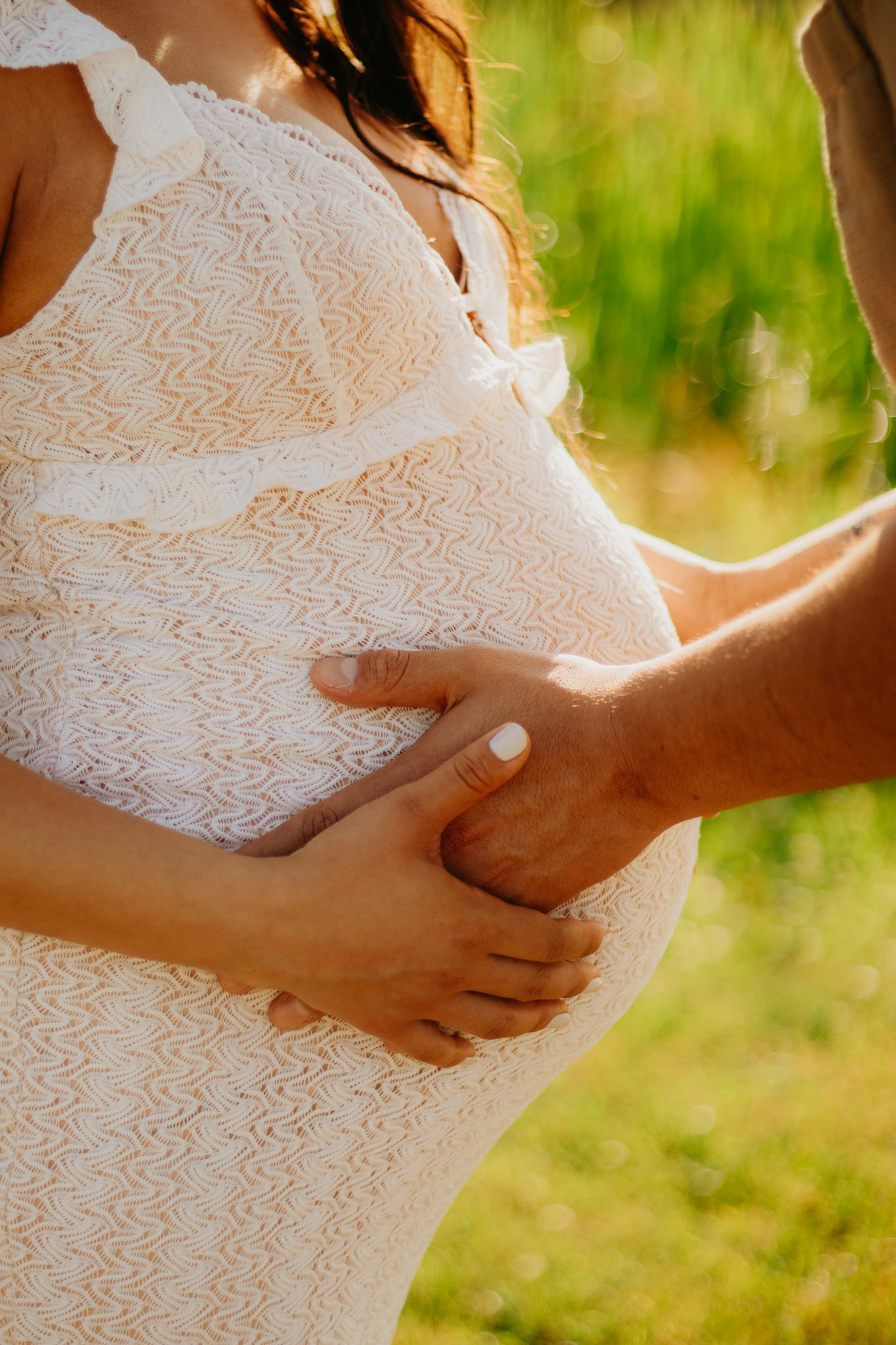 Detail shots focusing on hands gently placed on a pregnant belly in a white lace maternity dress.