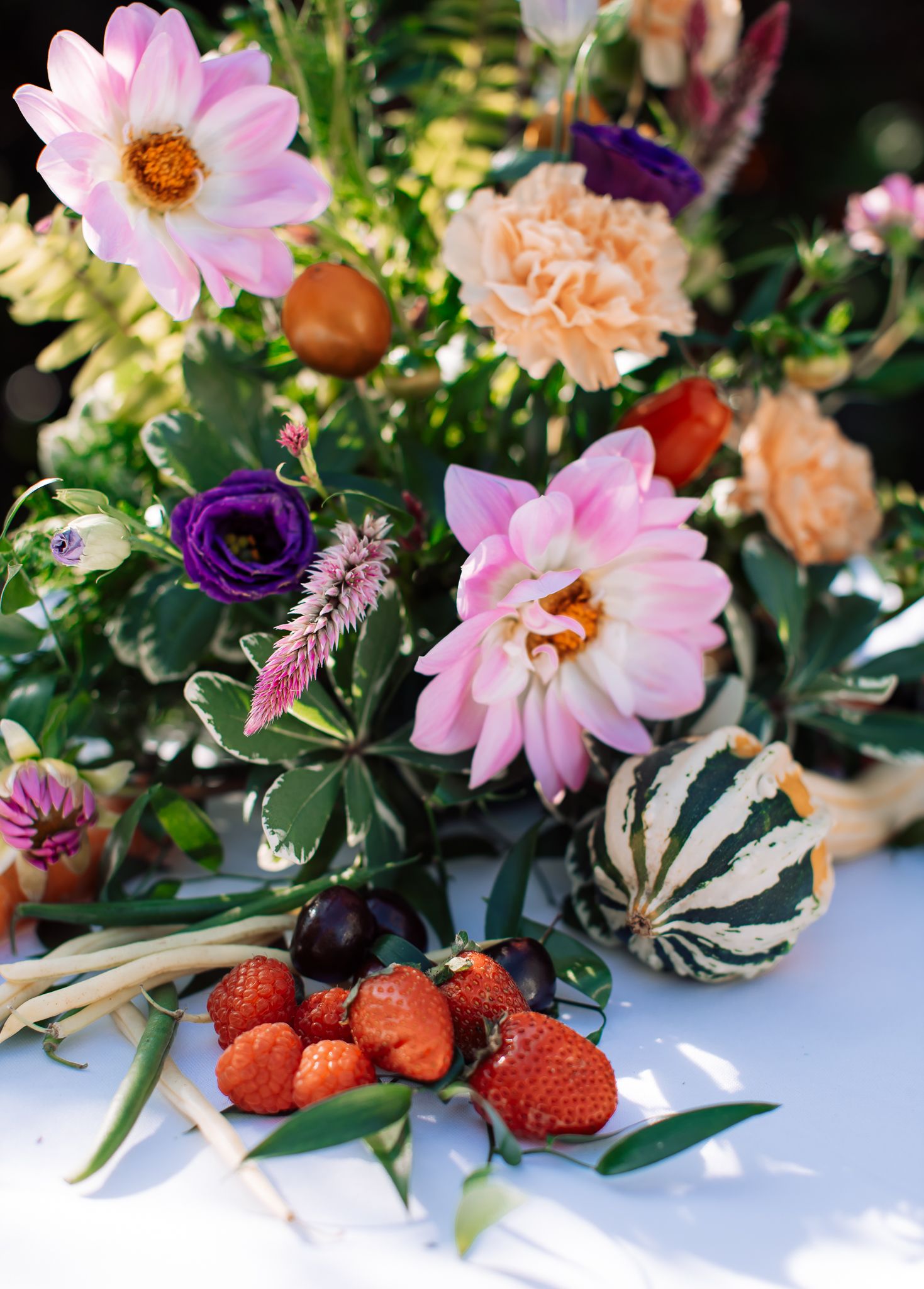 Colorful floral arrangement with pink dahlias, orange carnations, purple flowers, strawberries, and a striped gourd centerpiece.