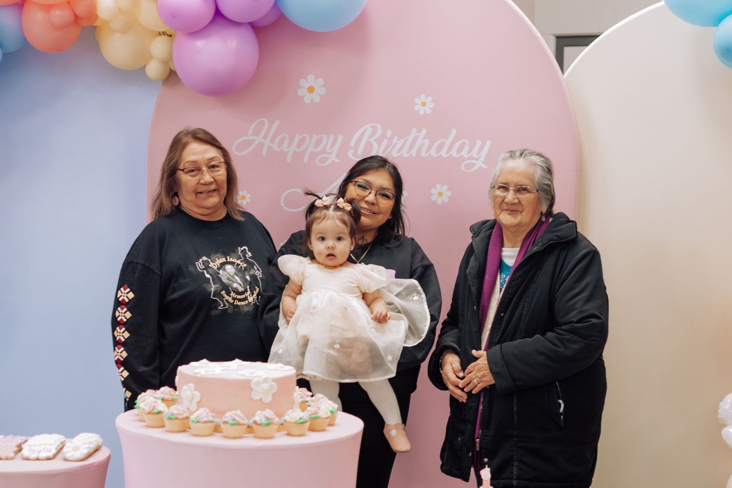 Three women and a baby celebrate a first birthday party with a pink cake and colorful balloon backdrop.