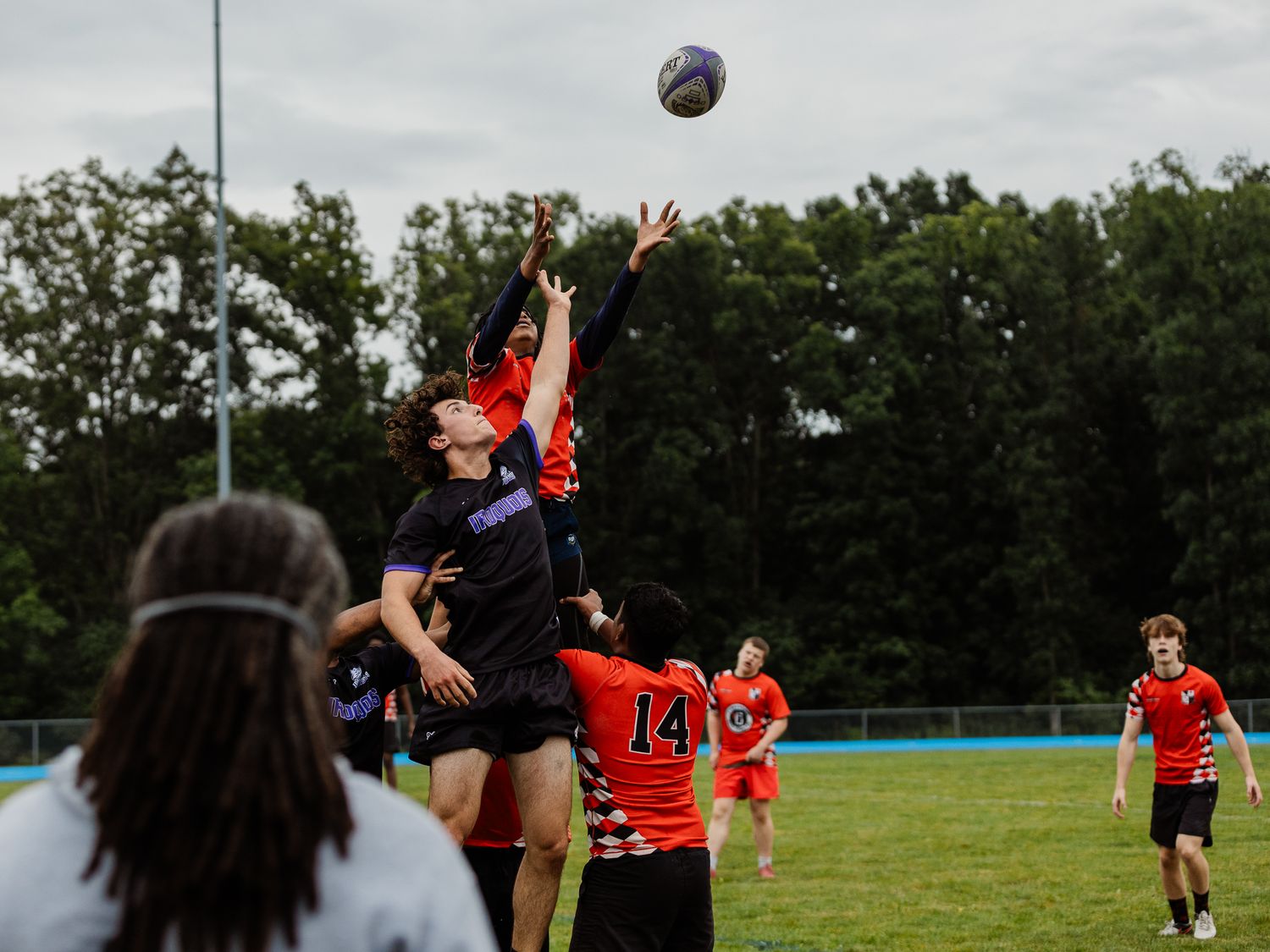 Players compete in a lineout during an outdoor rugby match on a green field with trees in the background.