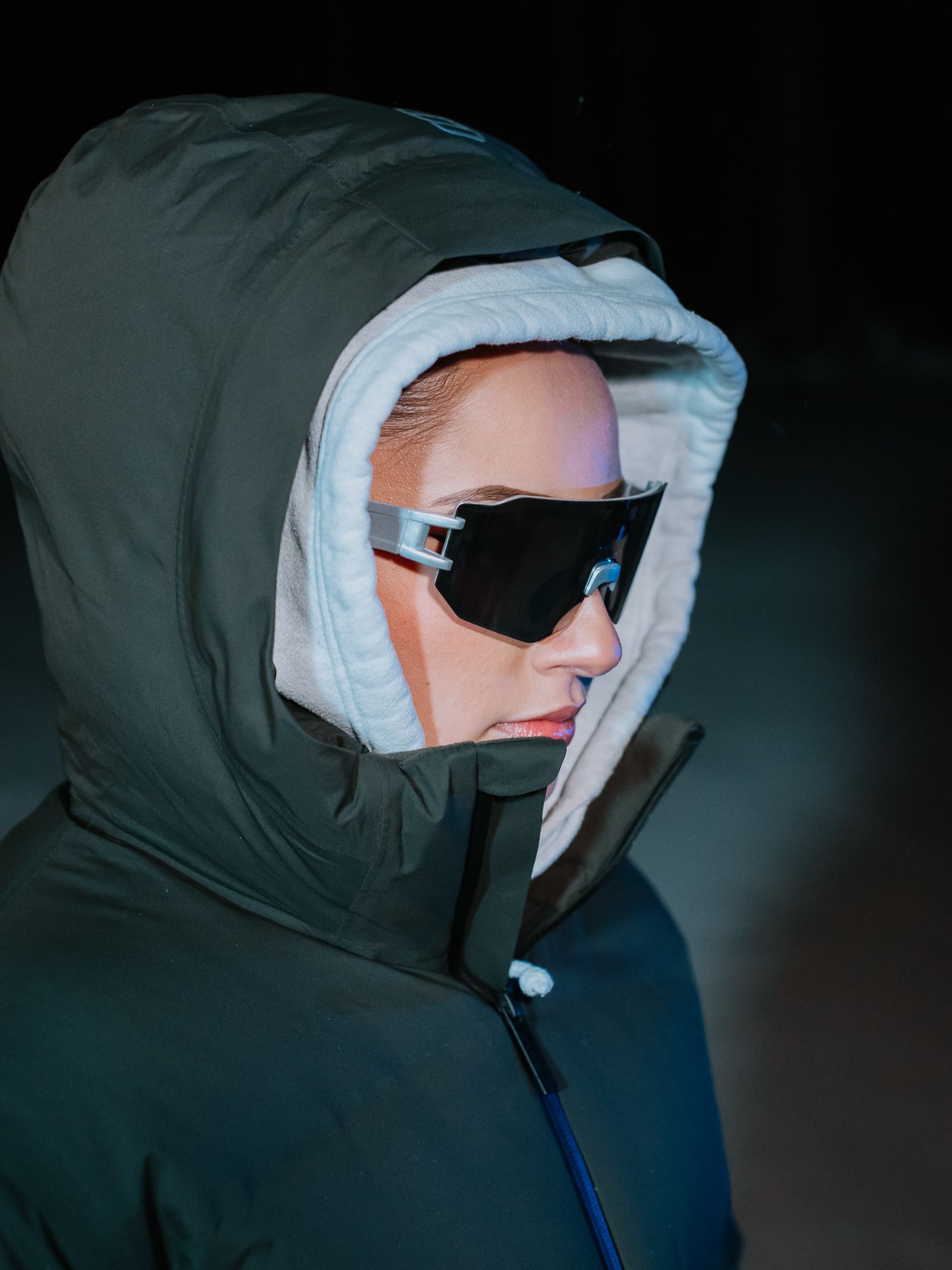 Person wearing dark hooded puffer jacket with white inner lining and black wraparound sunglasses against dark background.