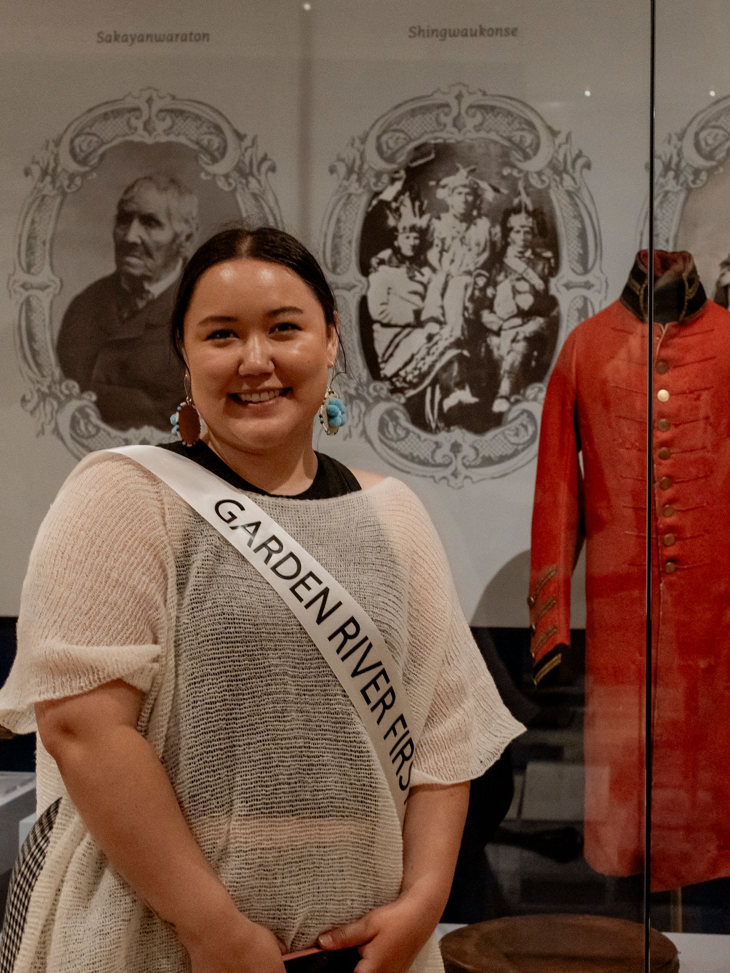 A smiling woman wearing a Garden River First Nation sash stands before historical Indigenous exhibits.
