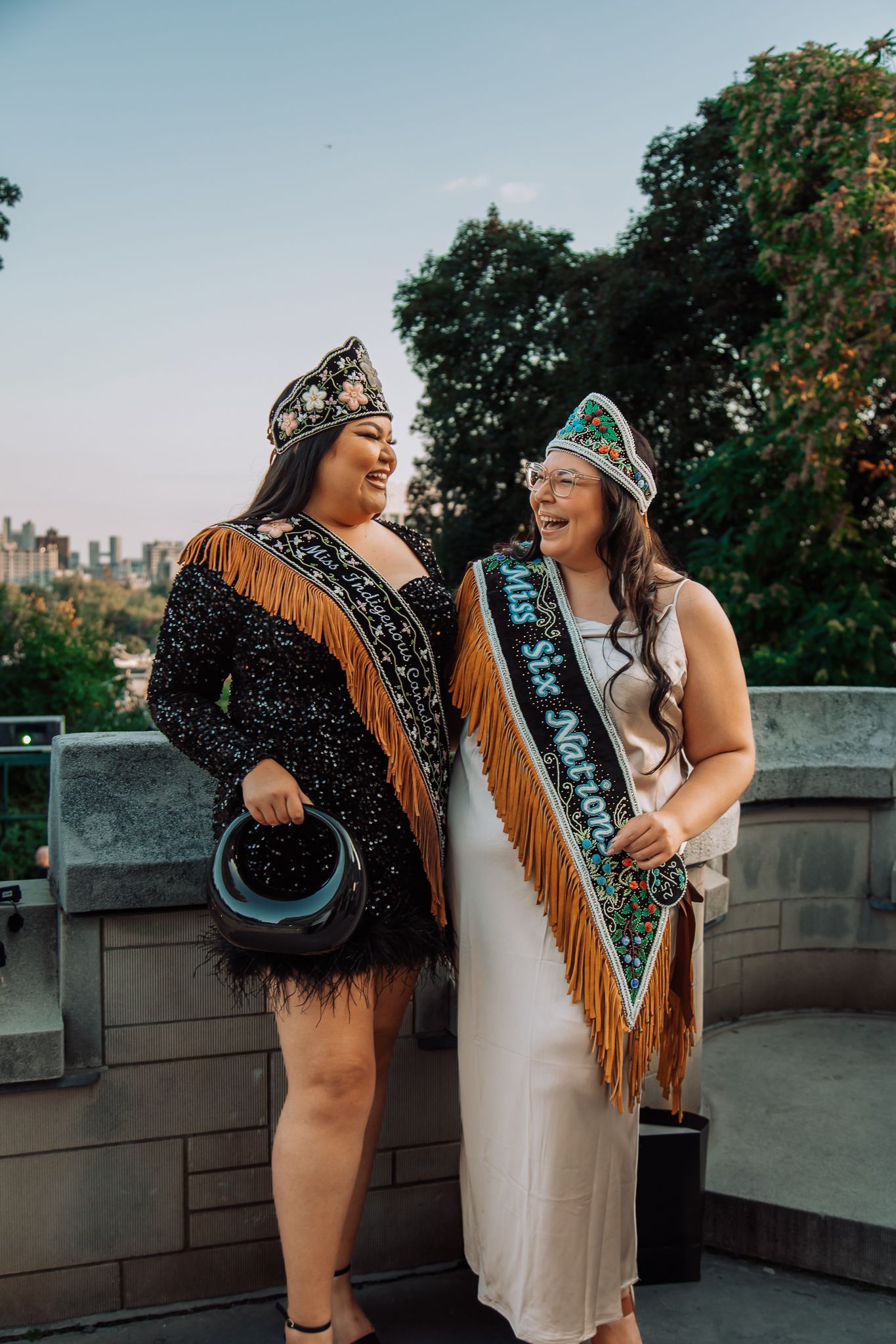 Two women in Indigenous regalia with fringed outfits and crowns, one wearing a Miss Six Nations sash, smiling outdoors.
