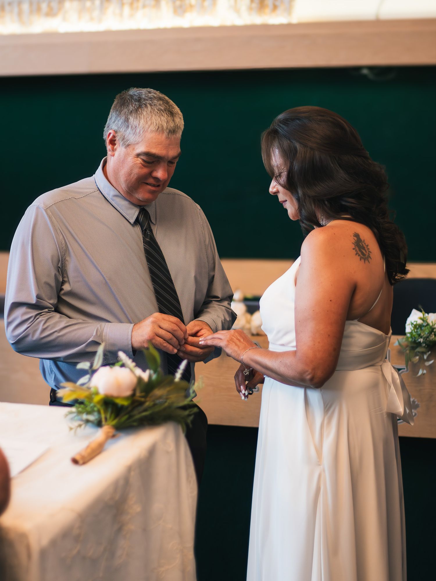 Groom placing wedding ring on brides finger during indoor ceremony, bride in white dress with floral bouquet nearby.