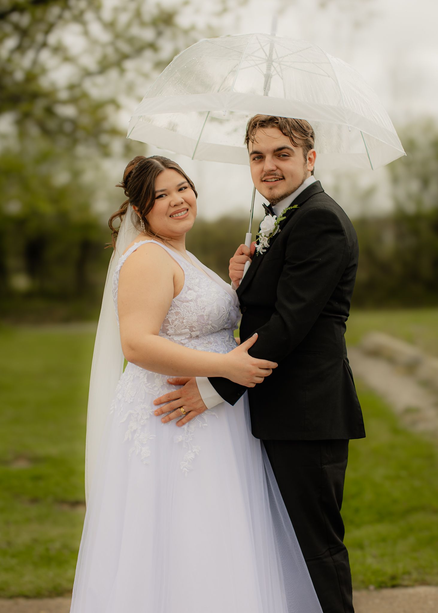Smiling bride and groom pose under clear umbrella on cloudy outdoor wedding day.