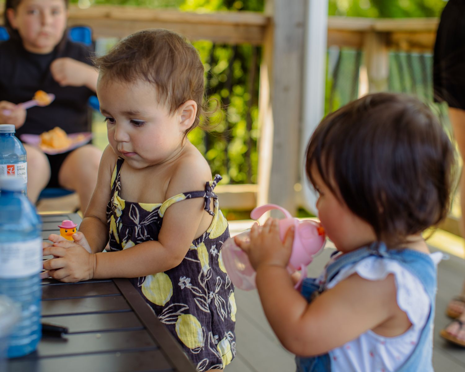 Two toddler girls sit at outdoor table sharing snacks and drinks at a summer party.