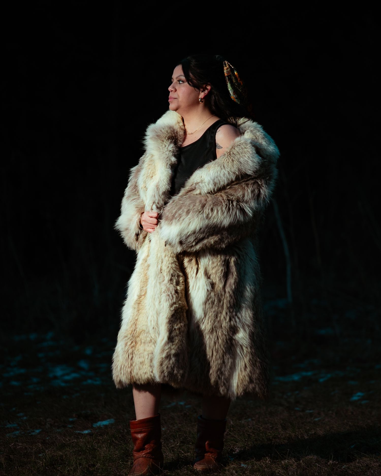 Woman in cream fur coat and brown boots standing in dark outdoor setting at night.