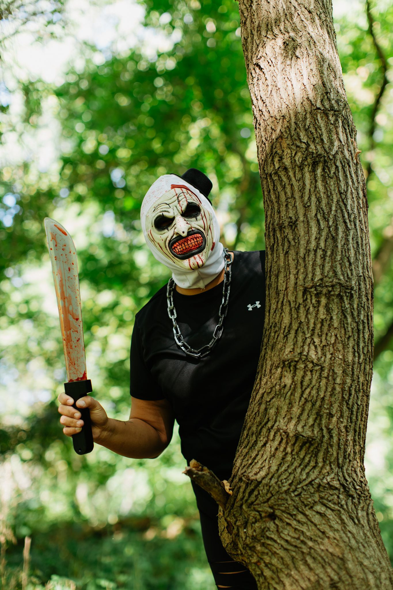 Person in a creepy clown mask and black shirt poses menacingly with a prop weapon behind a tree.