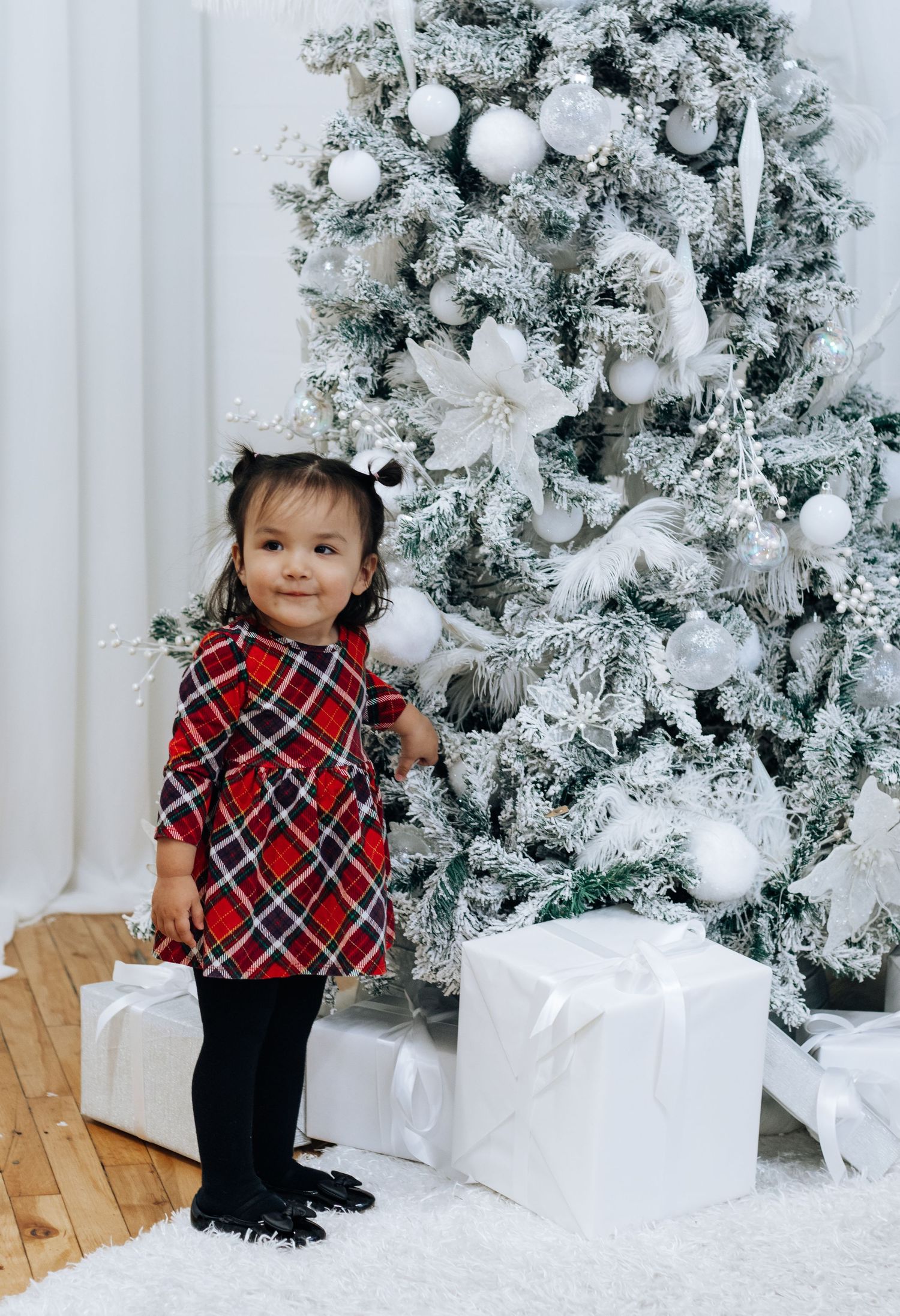Small child in plaid outfit standing by white flocked Christmas tree with wrapped presents.