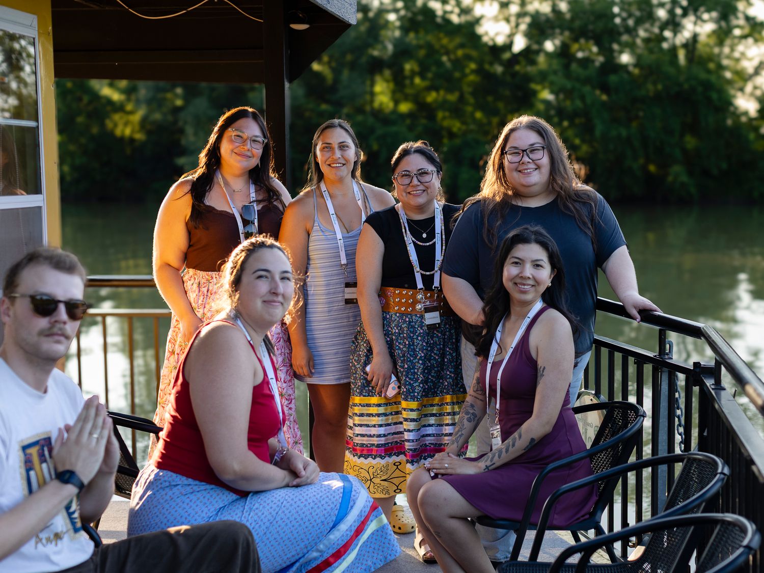 A diverse group of seven people posing together on a waterfront deck, smiling for a group photo outdoors.