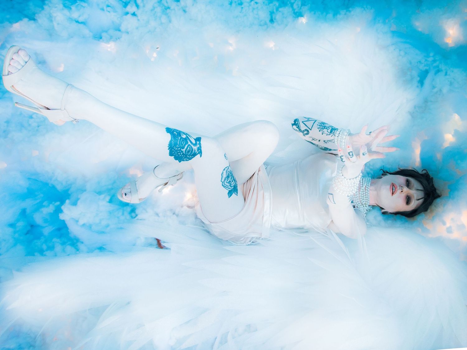 A person in white costume lies among blue and white clouds in a dreamy, ethereal fantasy photo shoot.