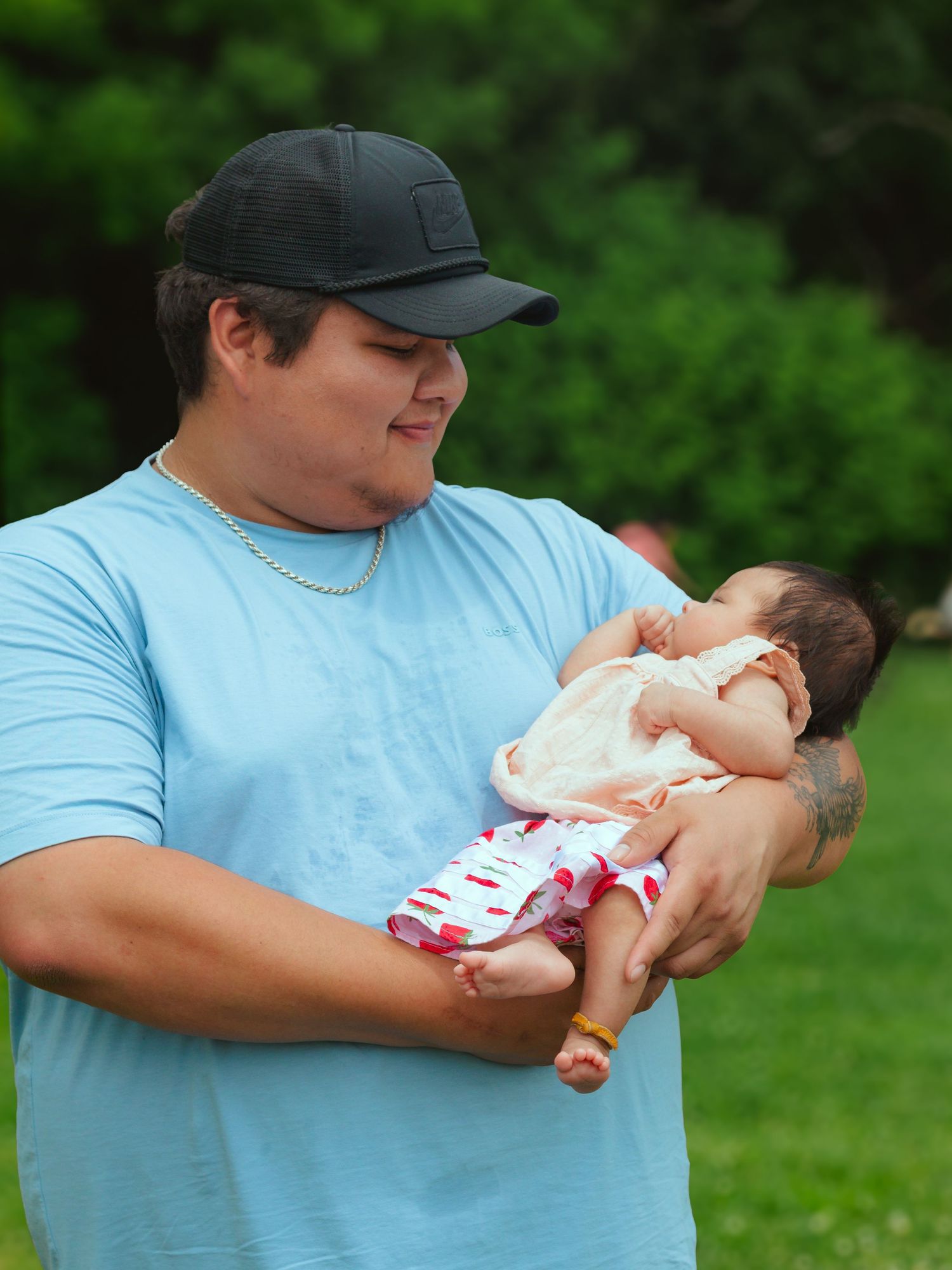 A young man in a blue shirt and black cap tenderly holds a newborn baby outdoors in a green park setting.