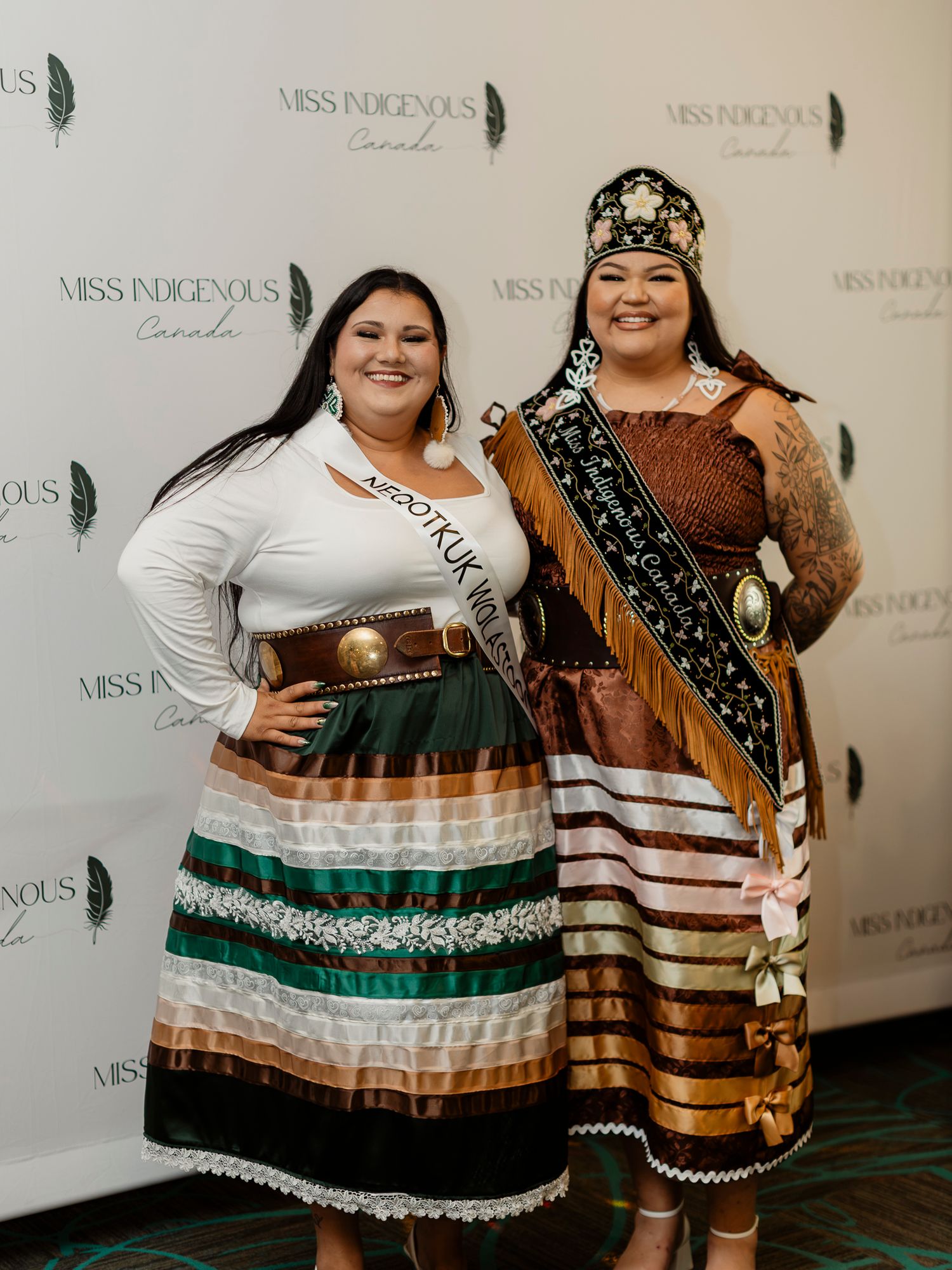 Two women in traditional Indigenous regalia pose at Miss Indigenous Canada pageant backdrop.