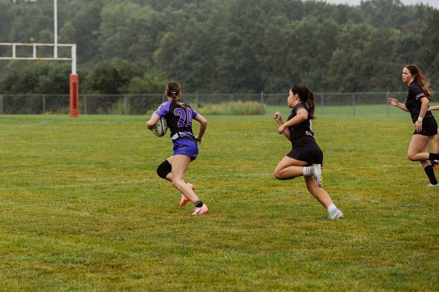 Female rugby players compete on a green grass field during a match, with one player carrying the ball while others pursue.