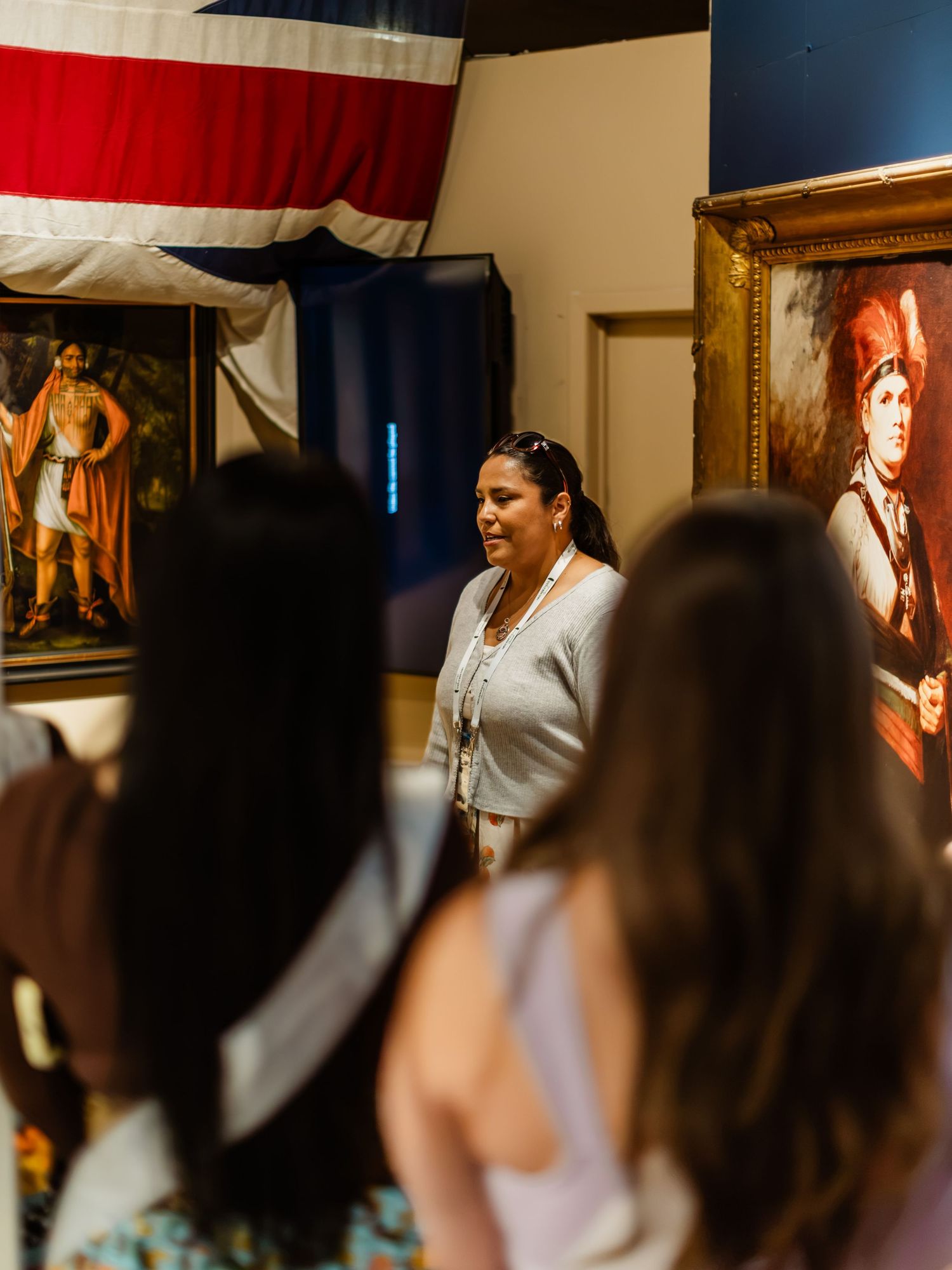 A museum guide presents paintings to a group of visitors in a gallery with historical portraits and a flag.