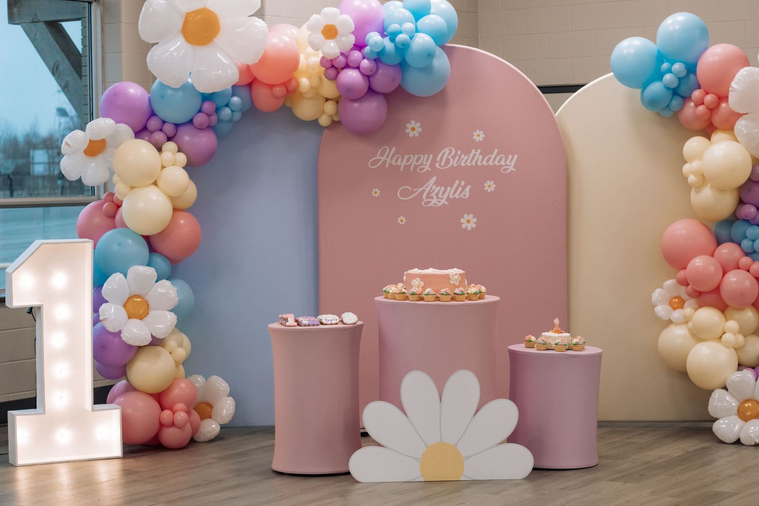 Pink and pastel balloon arch with daisy decorations and number one light display for birthday setup.