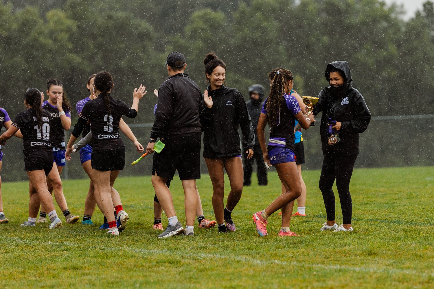 Female rugby players exchange high fives with coaches on a rainy sports field after a game.