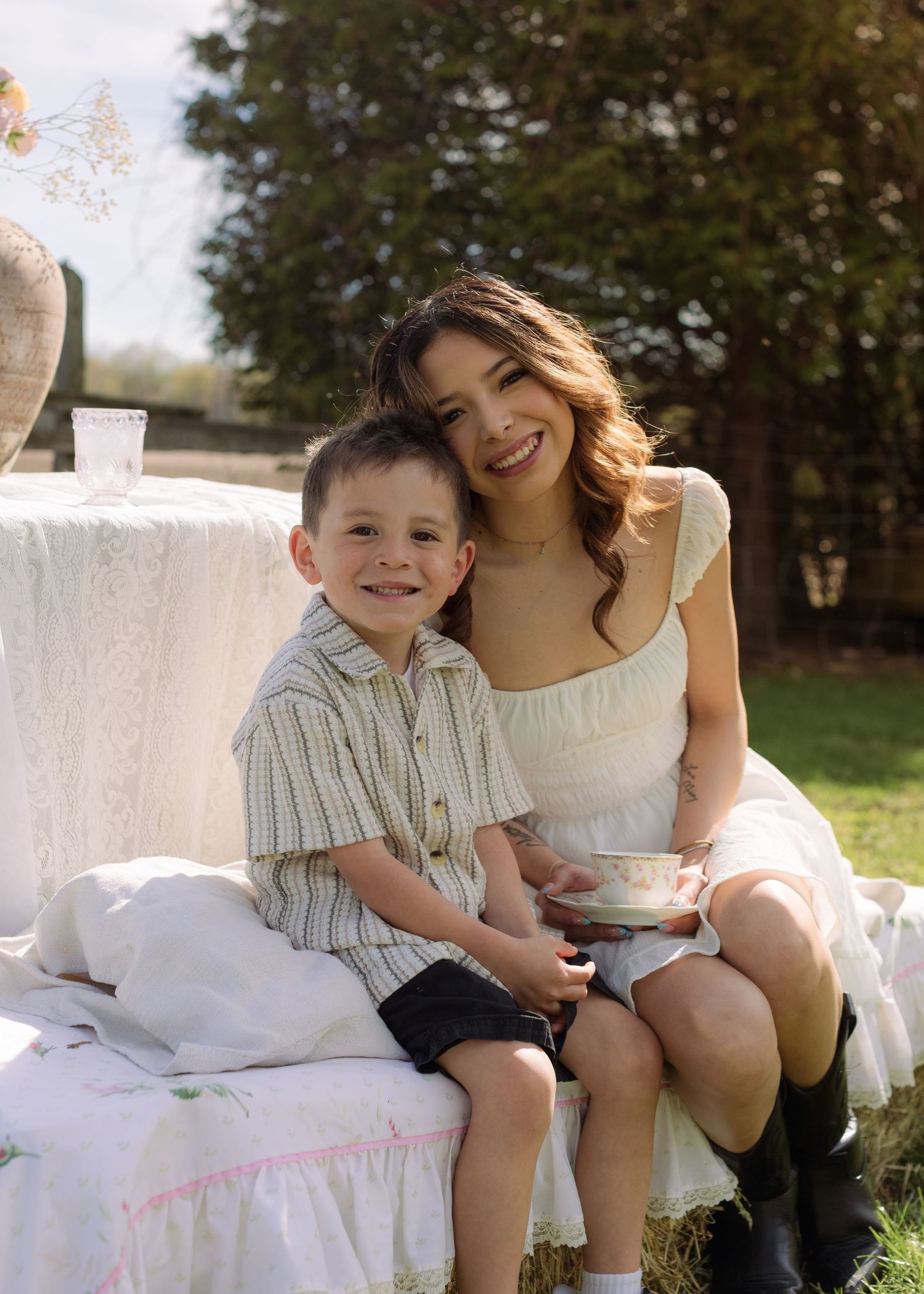 A smiling woman in a white dress holds a teacup beside a happy young boy at an outdoor tea party setting.
