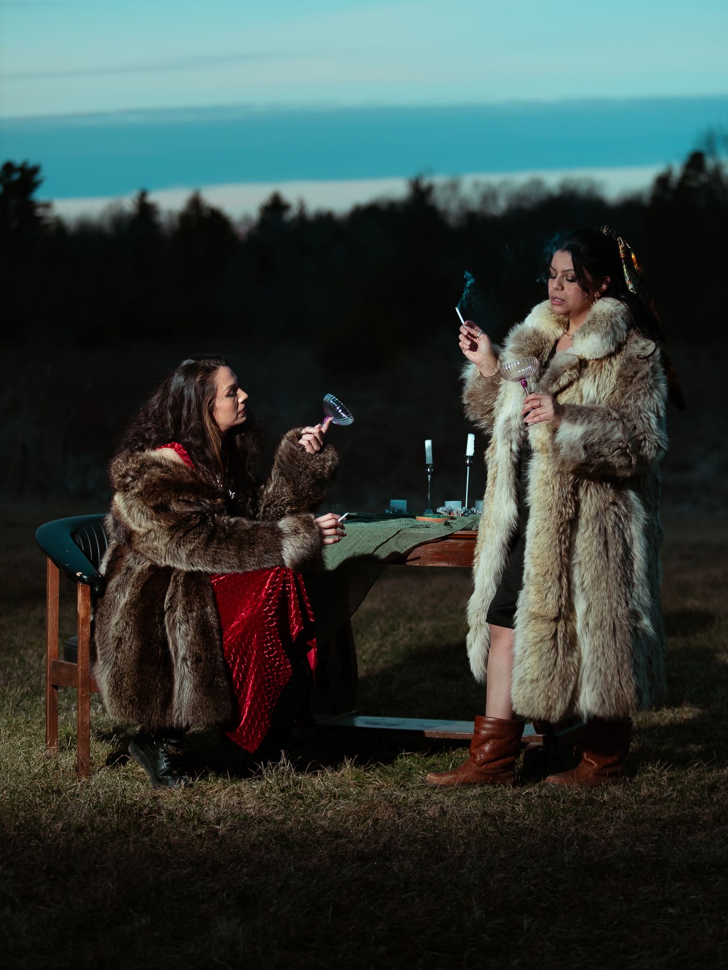 Two women in fur coats smoke at an outdoor candlelit table at dusk.