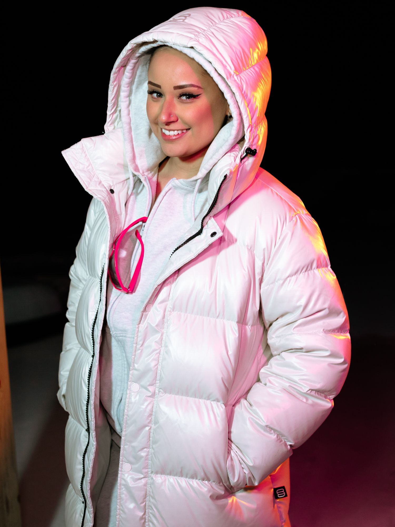 Smiling woman in shiny white hooded puffer coat illuminated by pink light against dark background.