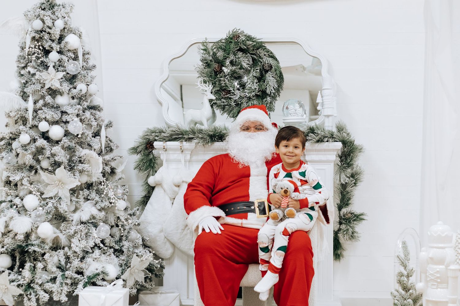 A child sits on Santa's lap in front of a decorated Christmas fireplace and flocked tree.