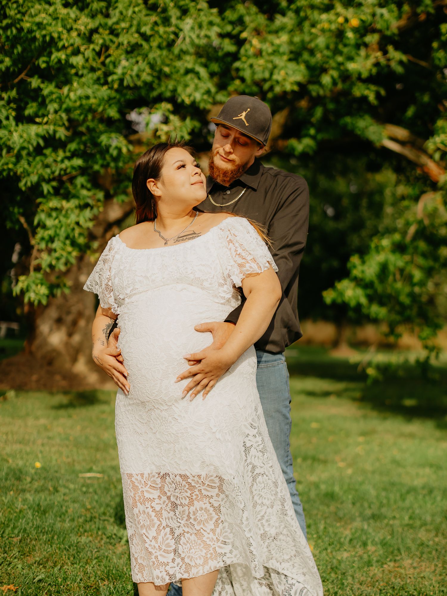 An intimate maternity portrait captured outdoors under green trees.