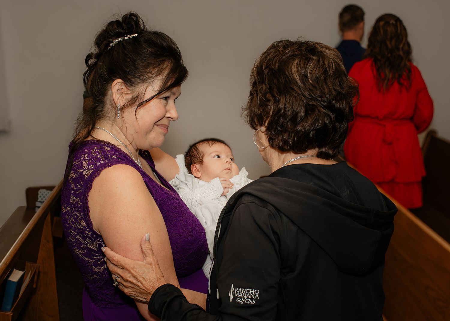 Woman in purple lace dress holds newborn baby in white christening gown at church ceremony.