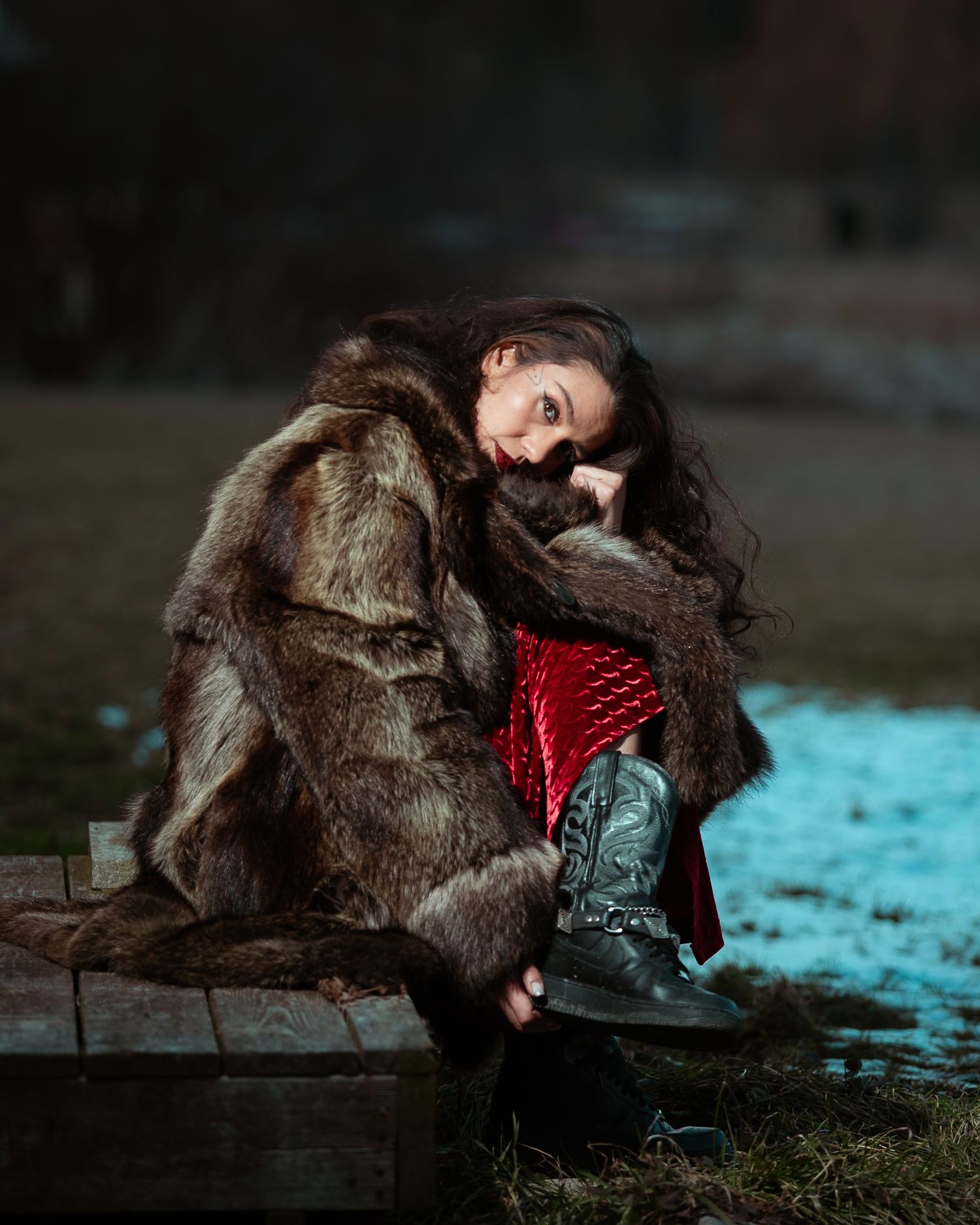 Woman in fur coat and red dress sits outdoors in winter light.