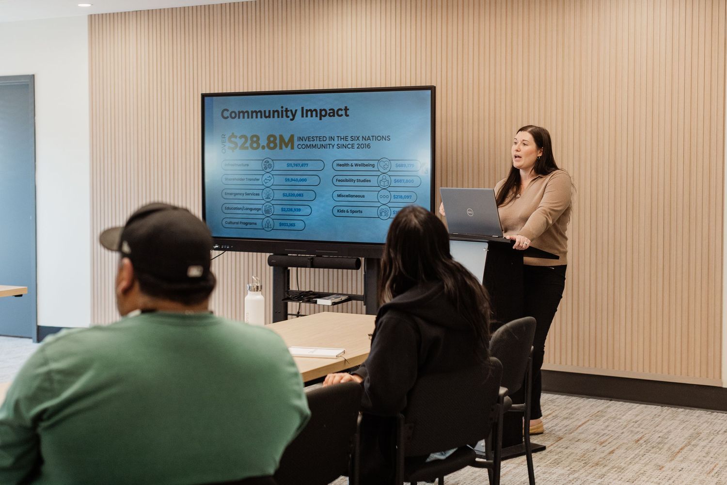 Woman presents Community Impact slide showing $38.8M to seated audience in modern conference room.