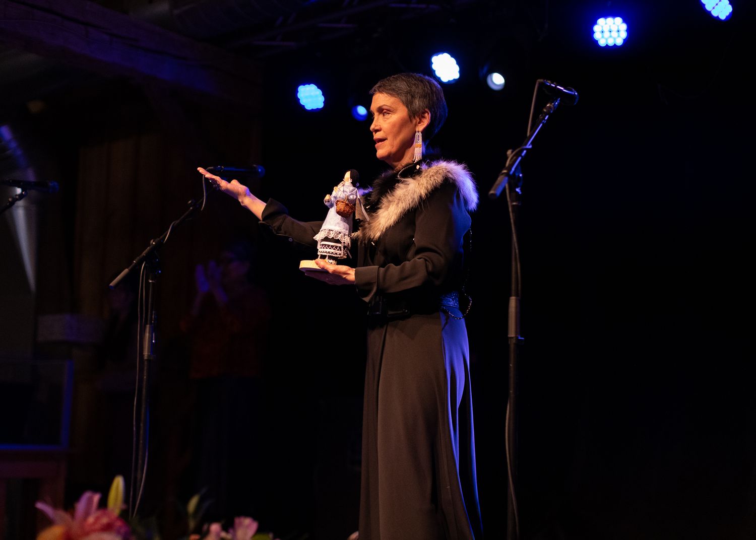 A woman in a black fur-trimmed dress accepts an award on a dimly lit performance stage.