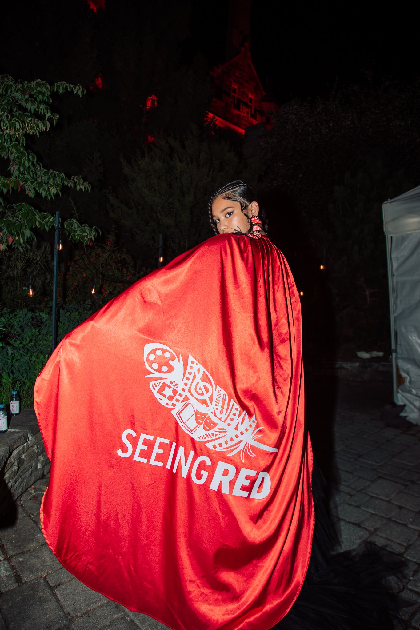 Woman dramatically posing in red satin cape reading Seeing Red with Indigenous artwork design.