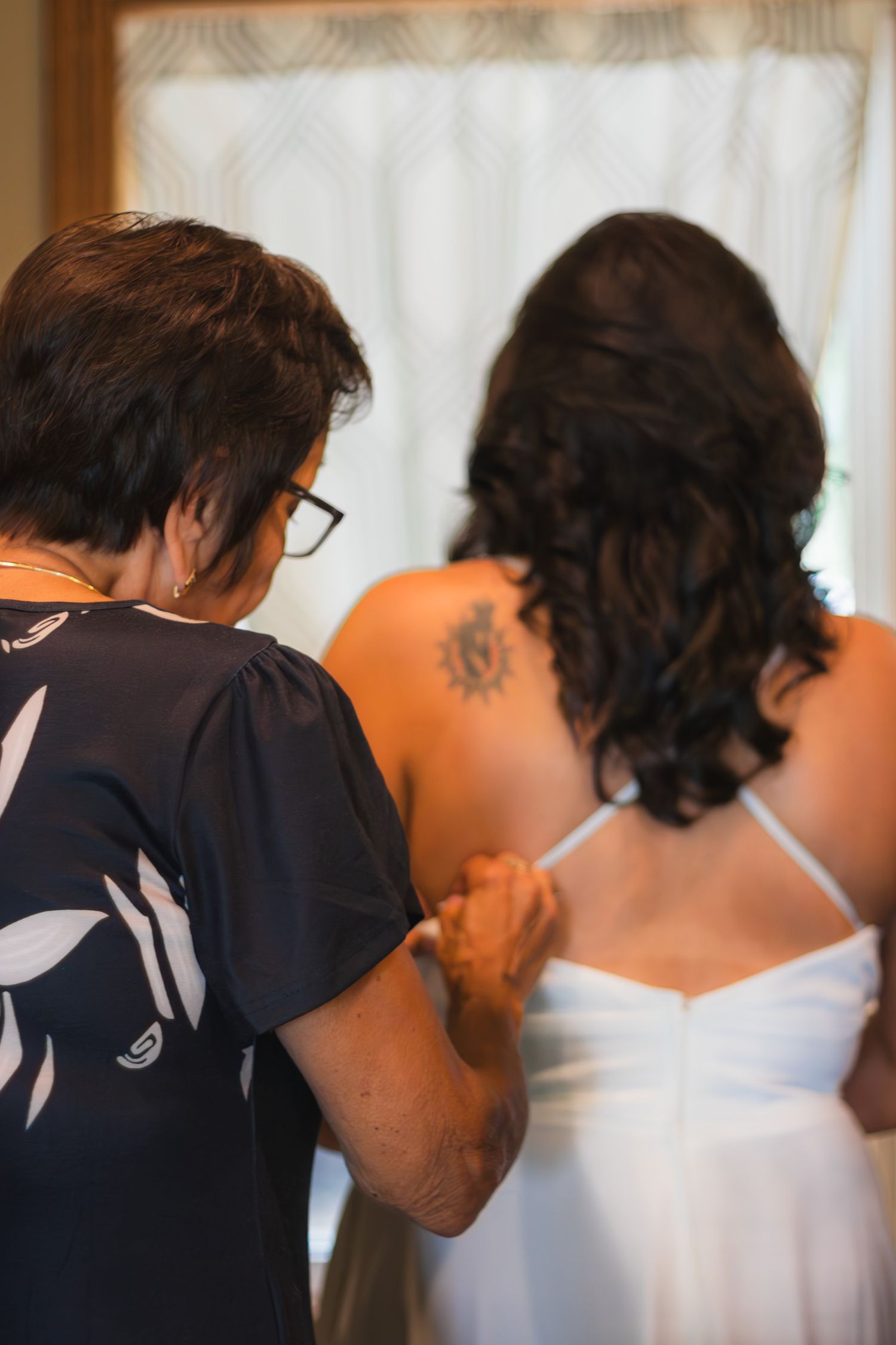 Older woman helps bride with white wedding dress zipper, bride has sun shoulder tattoo.