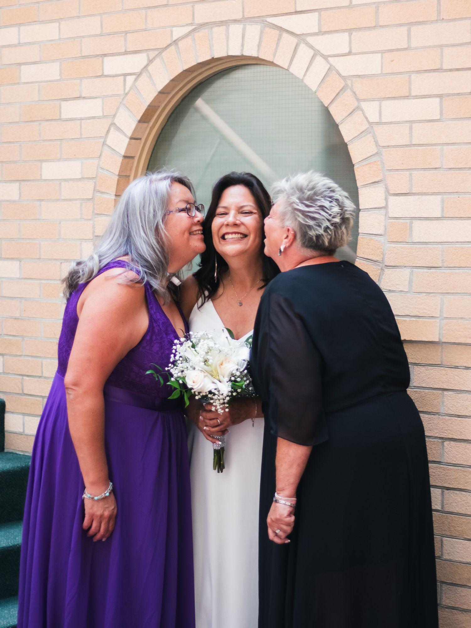 Bride in white gown holds bouquet while two women in purple and black dresses share a joyful moment outside a brick church.