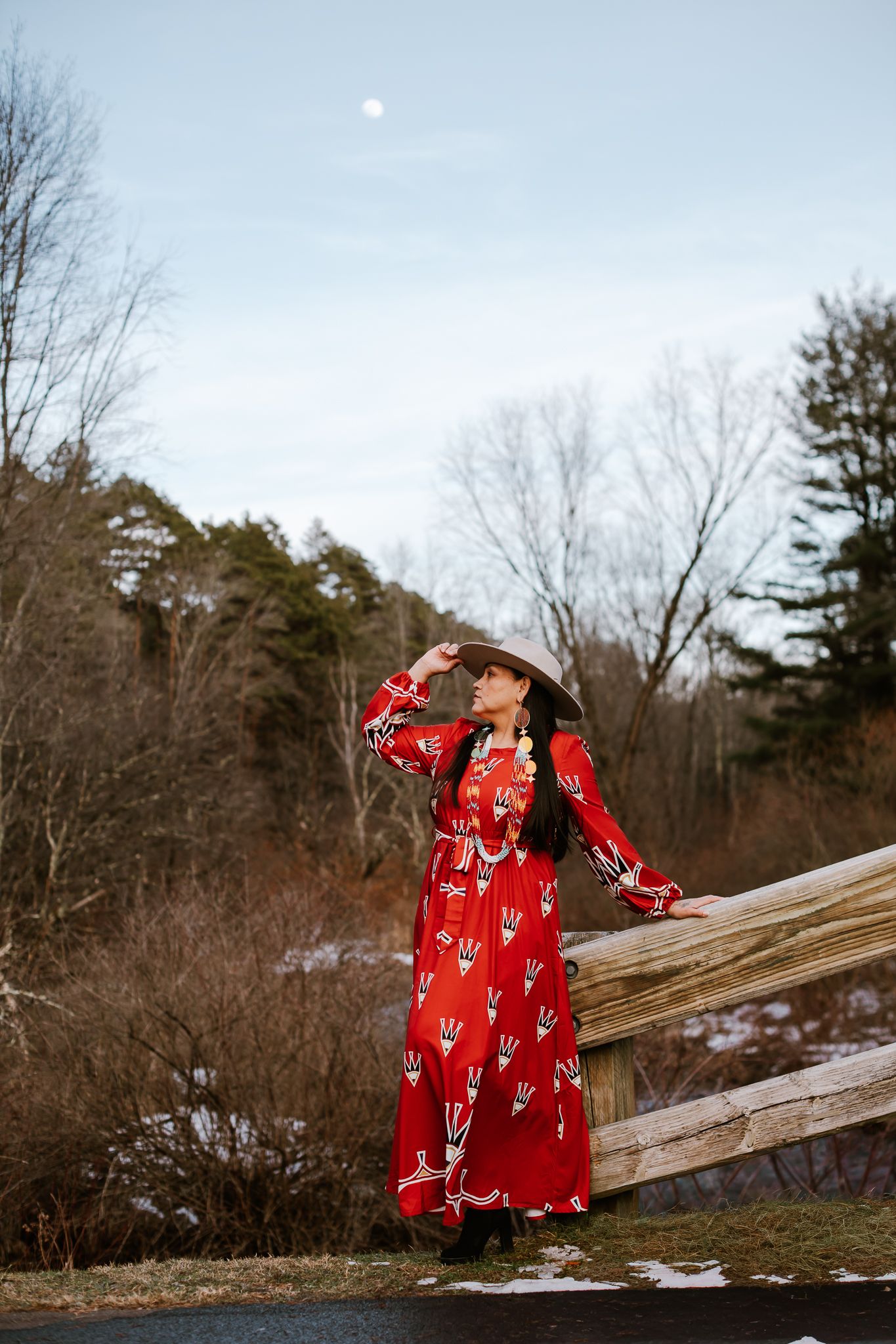 Woman in red patterned dress and cowboy hat poses by wooden fence in winter landscape with moon visible.